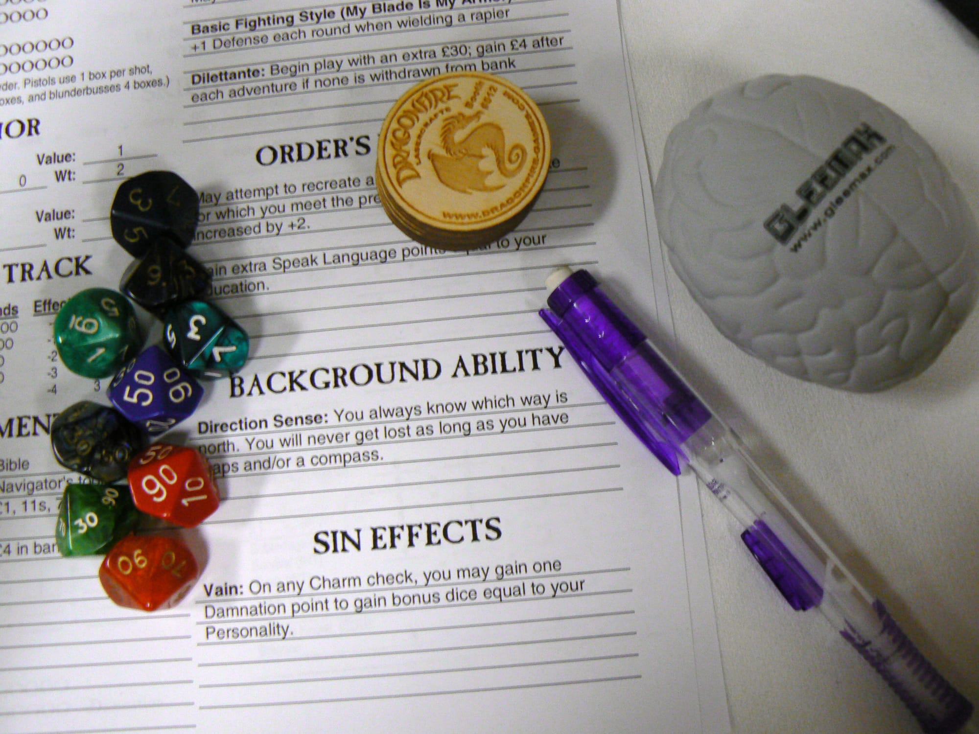 Several die, a pencil, some coins and a grey brain stress toy sit on top of a RPG charecters sheet.